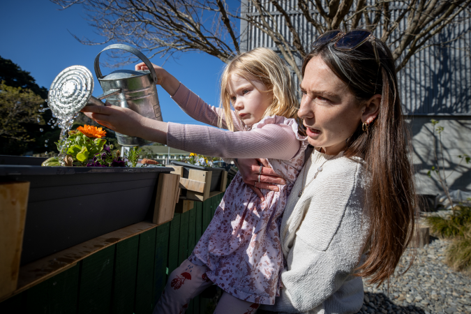 A kaiako and child outside. The kaiako is holding a child up to water a plant with a watering can.