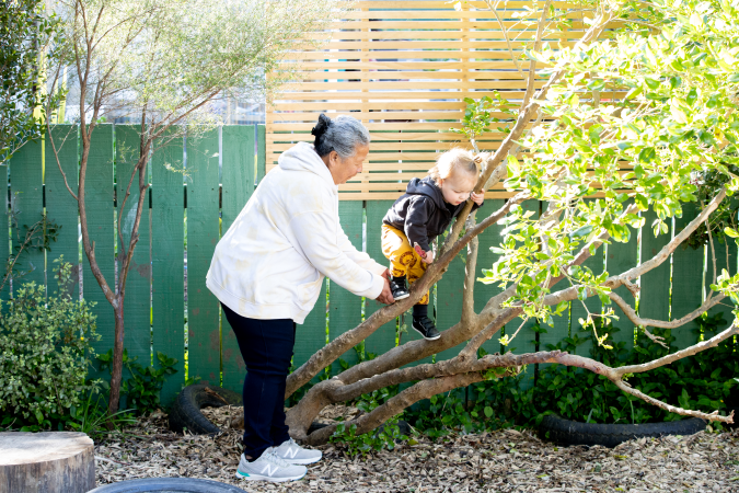 A toddler climbing the branch of a small tree outside with the support of a kaiako.