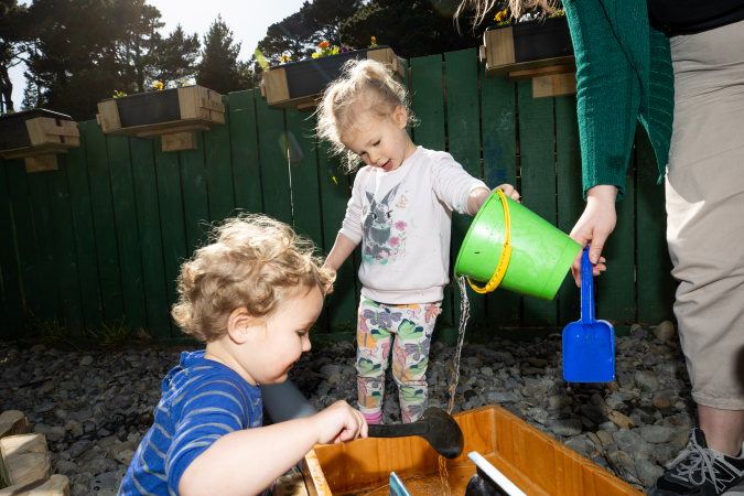 Young children and kaiako with a spade, spon and bucket pouring and measuring water in a wooden container.