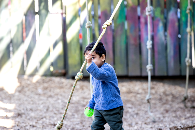 A child playing with a rope outside. A green ball is in their right hand.