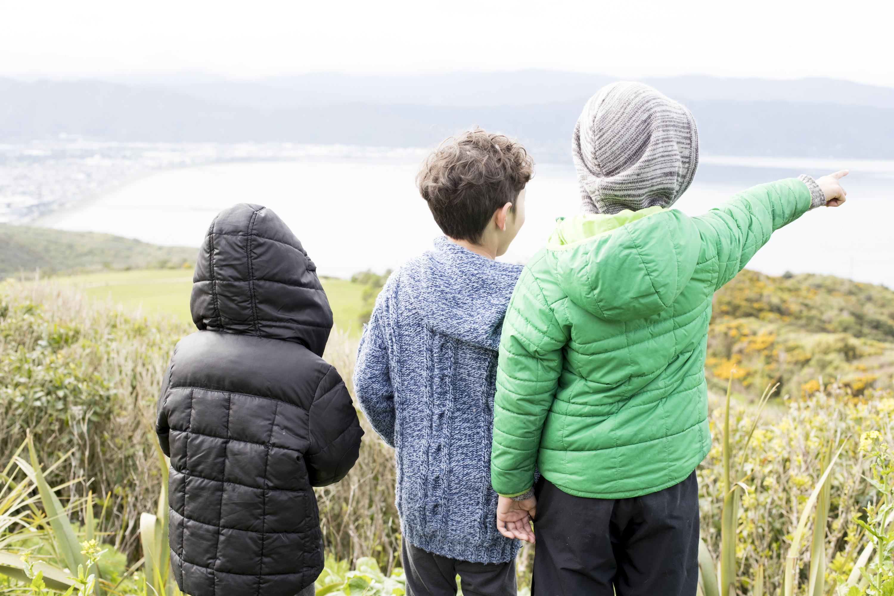 The back of three children in winter jackets, standing on a hill looking over a harbour. One child is pointing to the ocean. 