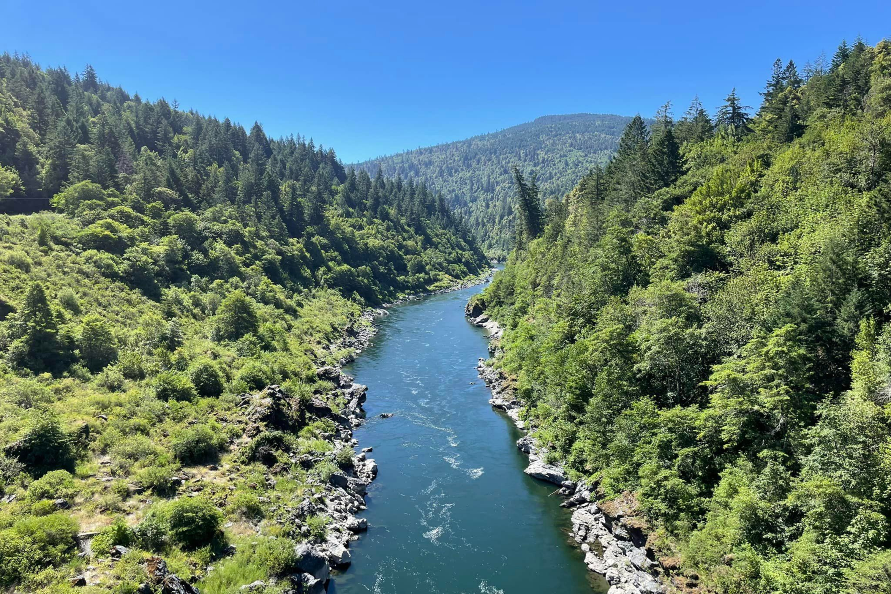 A blue stream flowing in the middle of two tree lined hills on a bright, sunny day. 