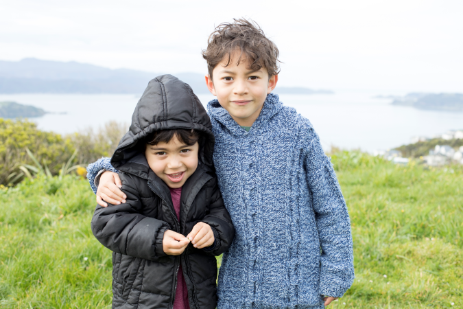 Two tamariki embracing, smiling and looking at the camera outdoors on a grassy hillside overlooking the sea.