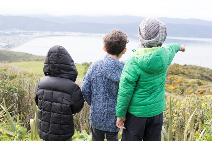 Three tamariki on a hillside looking over the horizon at the sea below.