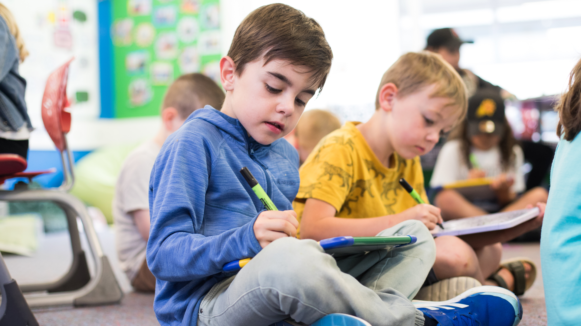 Two students in years one or two are sitting on the mat in a classroom each reading a book.