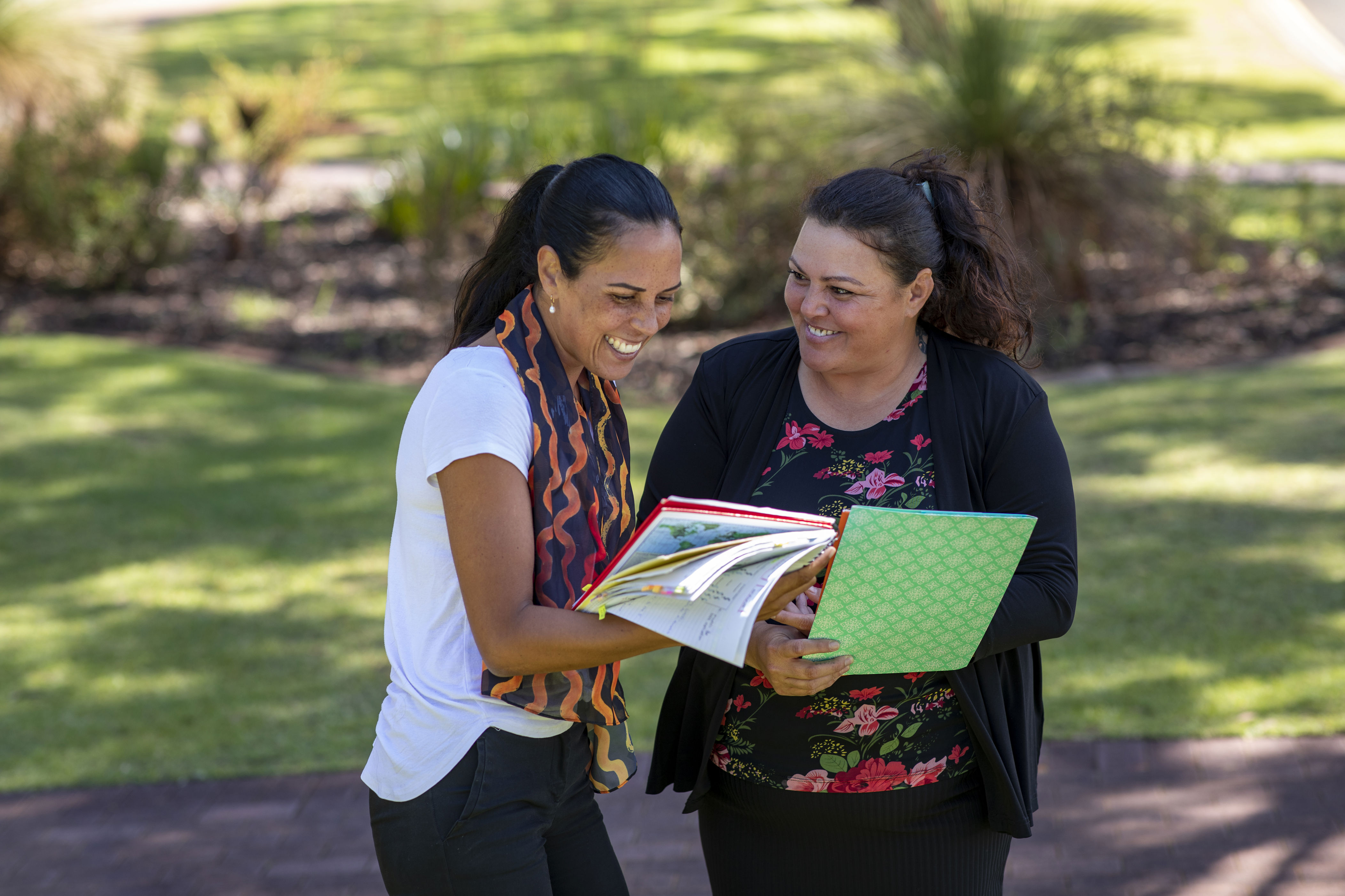 Two teachers speaking with each other outside, holding student workbooks. 