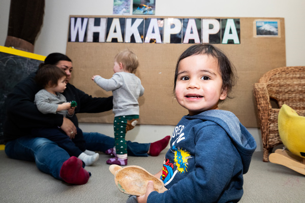 Tamariki smilling with a kaiako sitting with two other tamariki in the background and a sign with whakapapa on it