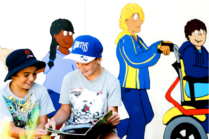 Two students sitting in front of a mural showing a person assisting a wheelchair user.