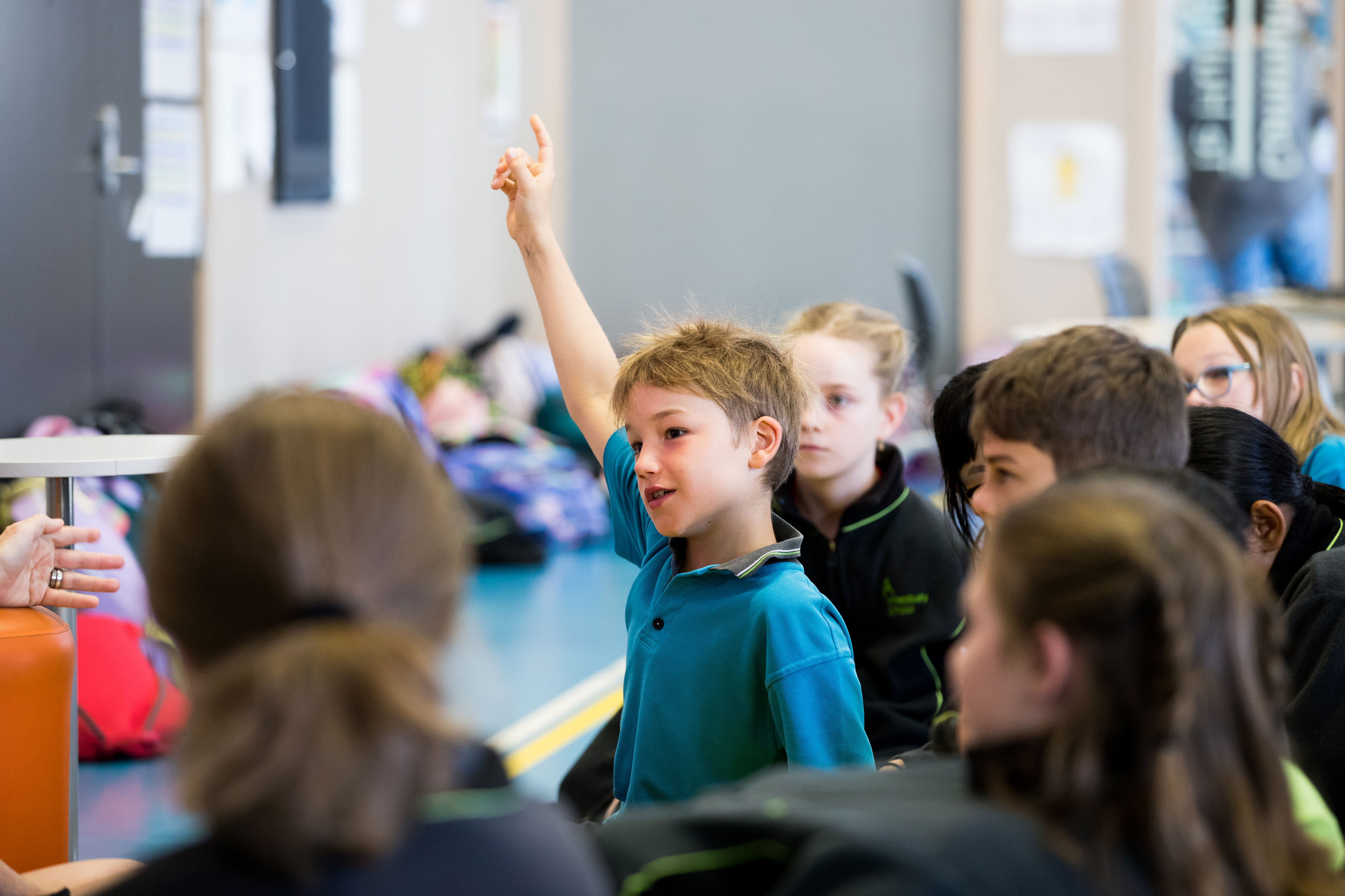 A primary school aged child raises their hand enthusiastically in a classroom setting, surrounded by other students.